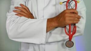 Una foto de un profesional de la salud con una bata blanca de laboratorio con los brazos cruzados sosteniendo un estetoscopio.